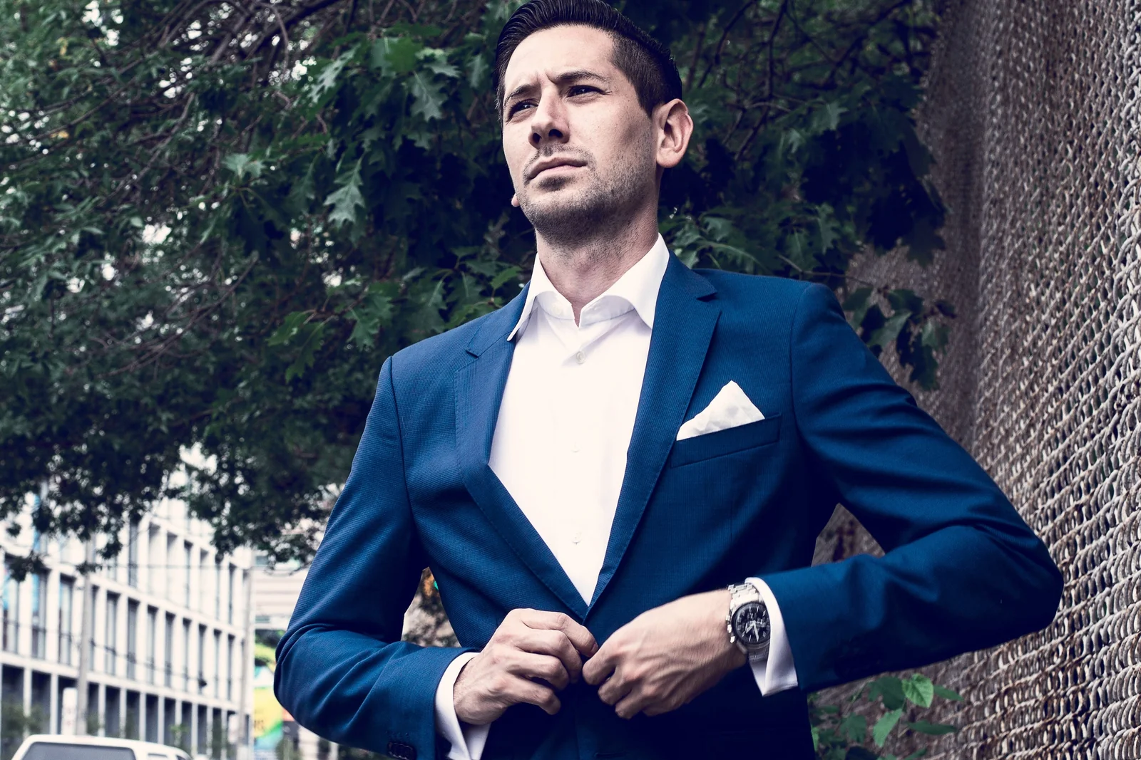 Man in a tailored navy suit and white shirt, buttoning his jacket on a city street